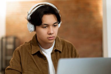 E-öğrenme kavramı. Kablosuz kulaklıklı genç Japon adamın portresi dizüstü bilgisayara bakıyor, çevrimiçi dersler, dersler ya da kapalı masa başında oturan sınıfları izliyor. Çocuk evde online çalışıyor.