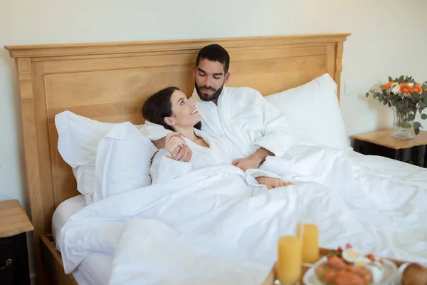 新婚旅行の喜び ホテルのベッドルームの屋内での朝食中にお互いに抱き合うカップルを愛しています 若い夫と妻が朝の高級スイートで寝そべっている — ストック写真