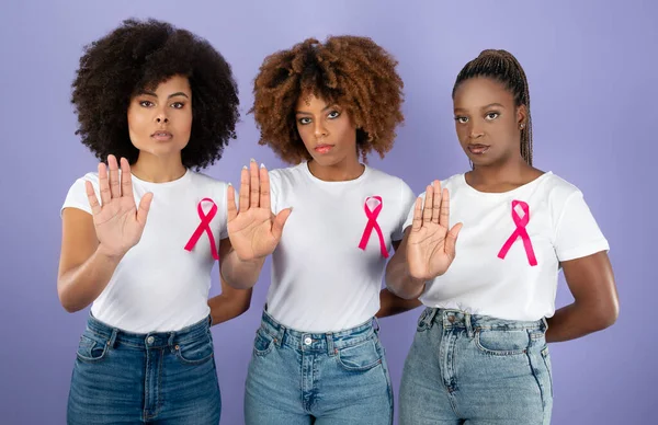 Oncology Awareness. Three Determined Black Women In T-Shirts With Pink Breast Cancer Ribbons Gesturing Stop Standing Over Purple Background, Looking At Camera. Studio Shot