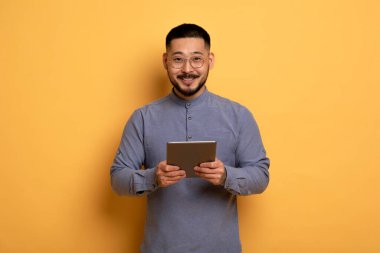 Yakışıklı Asyalı genç adam dijital tableti elinde tutuyor ve kameraya gülümsüyor, neşeli iş adamı, sarı stüdyonun arkasında dururken modern Gadget kullanıyor,