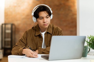 Çevrimiçi Çalışma. Kulaklıklı Asyalı genç adam evdeki bilgisayar dersinde bilgi yazıyor. Webinar 'a katılan zeki bir öğrenci, derslere kapalı alanda katılıyor.
