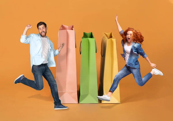 容易购物 季节性销售 身着休闲装 情绪化快乐的年轻夫妇们跳到一个色彩艳丽的购物袋旁边 摆弄着姿势 鬼鬼祟祟的 拼凑在一起 — 图库照片