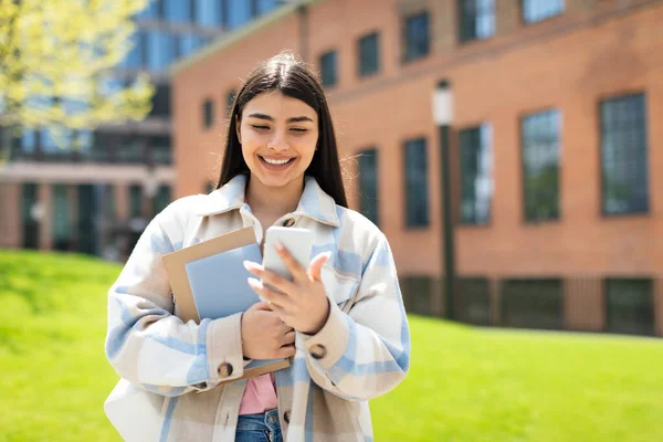 授業の後 大学のキャンパスを歩いて 屋外に立ってスマートフォンやワークブックを持つ陽気なヒスパニック系の女性の学生 友達との幸せな女性のチャットや笑顔 — ストック写真