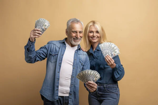 Wealthy rich happy elderly man and woman beautiful retired couple posing with cash money dollar banknotes isolated on beige background, get cashback, showing their savings or lottery winners