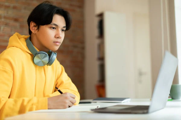 电子学习概念 聪明的亚洲少年在家里上网络课时 看着笔记本电脑在笔记本上写字 忙着参加在线讲座的学生坐在课桌前 — 图库照片