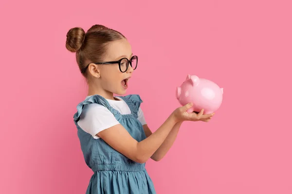 豚の銀行を保持している眼鏡で興奮した女の子の肖像 驚きのPreteen女性の子供は貯金や経済を楽しんで ピンクのスタジオの背景に隔離されたポーズ スペースをコピー — ストック写真