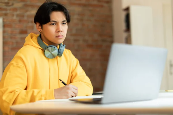远程学习概念 厌倦了在笔记本电脑上在线授课的亚洲少年 坐在家里听网上讲座时做笔记 专心致志的家伙在网上写作业 — 图库照片