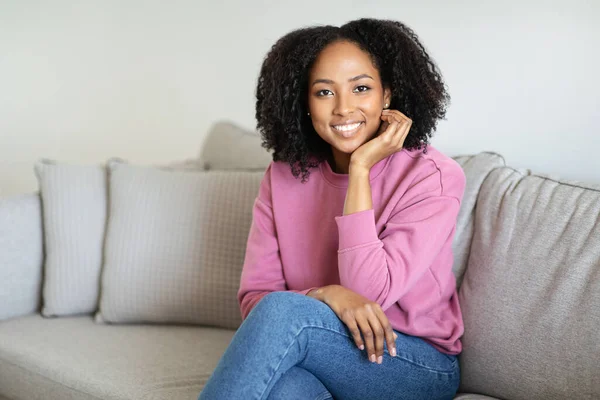 stock image Cheerful millennial black woman relaxing, enjoy free time, weekend, sit on sofa in minimalist living room interior. Housewife rest at home alone, ad and offer, good mood, lifestyle