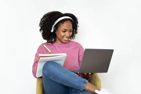 ワイヤレスヘッドフォンの陽気な千年の黒人女性は アームチェアに座って ノートパソコンでビデオを見て 白い壁の背景に注意を払っています 広告やオファー 自宅でガジェットとの作業 — ストック写真
