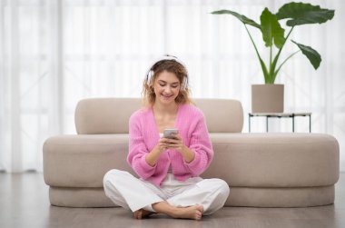 Gülümseyen Kadın Müzik Yayınları Servisi 'ne bakıyor ya da evdeki akıllı telefondan mesaj yolluyor, Oturma odasında yerde cep telefonuyla dinlenen kablosuz kulaklık takan mutlu genç bayan,