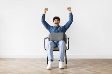 Neşeli haberler. Duygusal Ortadoğulu serbest çalışan bir adam dizüstü bilgisayarla koltukta oturuyor, online galibiyeti kutluyor, evet jestleri yapıyor ve beyaz duvarın üzerinde duran kameraya bakıyor. Şans ve Başarı