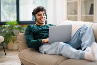Çevrimiçi Eğlence. Kablosuz kulaklıklı gülümseyen Arap adam evde dizüstü bilgisayar kullanıyor, koltukta dinleniyor bilgisayarda film seyrediyor ya da müzik dinliyor. Serbest Çalışan Uzaktan İşlerin tadını Çıkartıyor