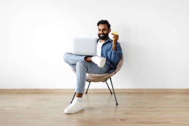 Evinde beyaz duvara yaslanmış modern bir koltukta dinlenirken internetten bilgisayar ve kredi kartıyla alışveriş yapan mutlu, genç bir Arap. Gülümseyen Adam İnternet Bilgisayarından Satın Almanın Zevkini Çıkartıyor