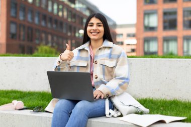 Mutlu İspanyol öğrenci bayan dışarıda dizüstü bilgisayarla oturuyor, işaret ediyor ve gülümsüyor, online eğitimi onaylıyor, kampüs parkında uzaktan ders alıyor. E-öğrenmeyi seviyorum.