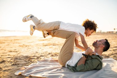 Neşeli genç babası ve oğlu birlikte eğleniyor, adam kumsalda ekoseyle yatıyor ve çocuğu ayaklarının üstünde büyütüyor, aile dışarıda vakit geçiriyor, yan gözle bakıyor.