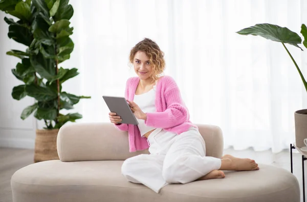余暇のための技術 自宅でデジタルタブレットを使用して幸せな若い女性 現代のガジェットとソファの上で休む千年紀の女性を笑顔 リビングルームのインテリアで快適なソファにくつろぎます — ストック写真