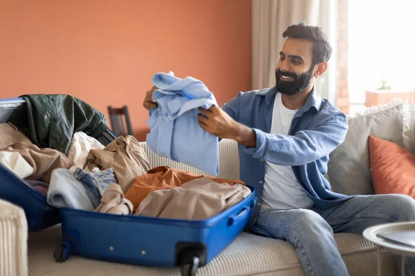 度假时尚 开朗的阿拉伯男子用旅行箱包装衣服 为旅行旅行准备行李 把他的衬衫放在家里的长椅上 时尚旅行的概念 — 图库照片