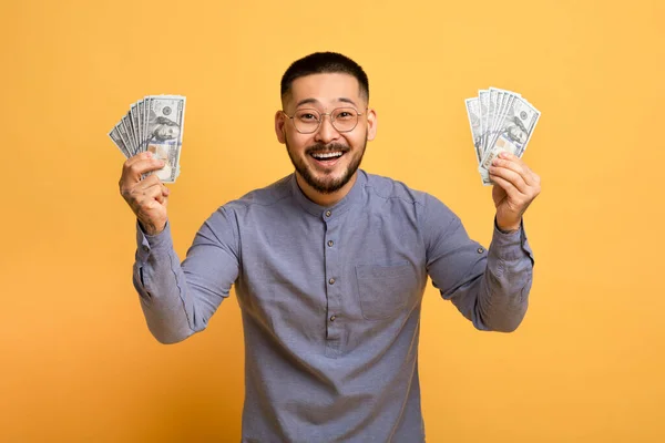 Lucky Winner. Excited Millennial Asian Man Holding Money Cash In Hands, Joyful Euphoric Young Guy With Dollar Fans Posing On Yellow Studio Background, Celebrating Big Profit Or Win, Copy Space