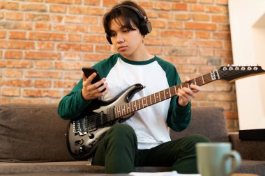 Asyalı ergen çocuk elektro gitar tutuyor Smartphone 'a bakıyor, internetteki Song akorlarını kontrol ediyor Modern Home Interior' da koltukta oturuyor. Kulaklıklı Adam Enstrüman çalmayı Öğreniyor