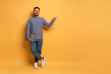 Güzel reklam. Gülümseyen Asyalı adam dizüstü bilgisayarını tutuyor ve sarı stüdyo arkaplanındaki boşluğu işaret ediyor, neşeli milenyum adamı mesajınız ya da tasarımınız için boş yer gösteriyor, tam uzunluk