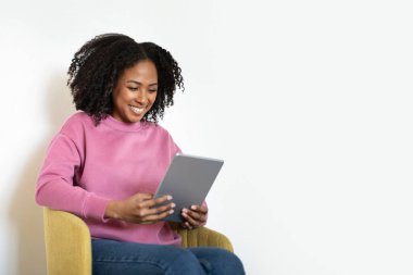 Gülen milenyum siyahi kadın, beyaz duvar arkasında, tablet üzerinde oturup sohbet ediyor. Çalışma, evde çalışma, sohbet, blog ve sosyal ağlar, video çağrısı