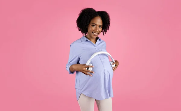 快乐的千年非洲裔美国卷曲孕妇 穿着休闲装 把无线耳机放在肚子里 享受未来的母性 等待婴儿 隔离在粉红的工作室背景下 — 图库照片