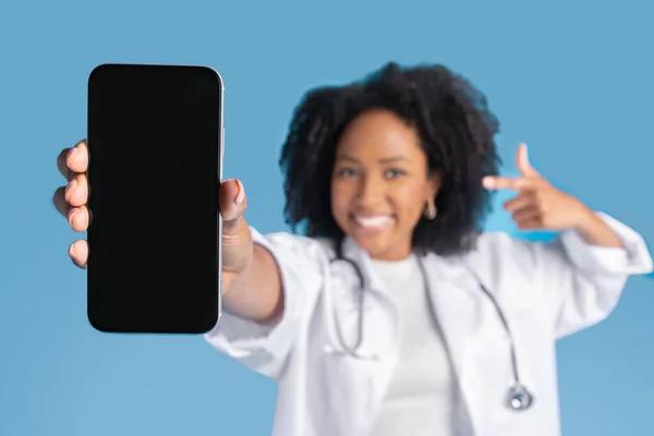 stock image Happy excited young back curly lady doctor in coat point finger at phone, focus on blank screen, isolated on blue background, studio, close up. Recommendation app, medicine, health care remotely