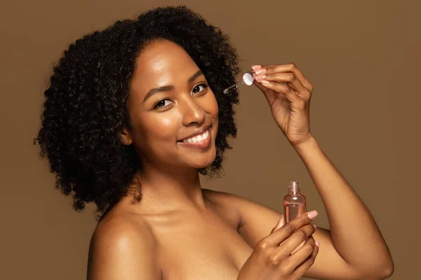 快乐漂亮的半裸黑人妇女拿着装有美容产品的瓶子 应用抗衰老血清 在棕色背景下分离 皮肤护理常规 面部化妆品 — 图库照片