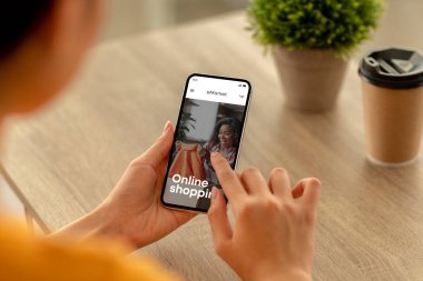 Genç bir kadın internetten alışveriş için cep telefonu kullanıyor, ahşap masaya oturmuş kahve alıyor, yeni kıyafetler arıyor ve seçiyor. Uygulama ya da web sitesi olan sipariş alımları, ekranda moda mağazası