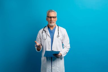Yakışıklı, gri saçlı, gözlüklü yaşlı adam elinde tıbbi dosya olan doktor, hastaya danışmanlık yaparken anamnezi alıyor, kameraya, jestlere, fotokopi alanına, mavi stüdyo geçmişine bakıyor.