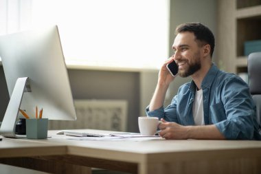 Milenyum işadamı ofisteki işyerinde cep telefonu ve kahveyle rahatlıyor, masasında oturmuş gülümseyen, cep telefonuyla konuşan, ailesiyle konuşan, iş yerinde mola veren...