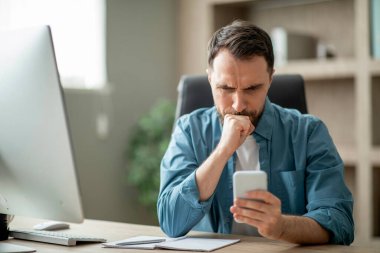 Stresli işadamı ofiste otururken akıllı telefon ekranına bakıyor, endişeli erkek girişimci SMS okuyor ve kaşlarını çatıyor, kötü haberler alıyor, dolandırıcılık mesajı alıyor, yakın çekim çekiyor.