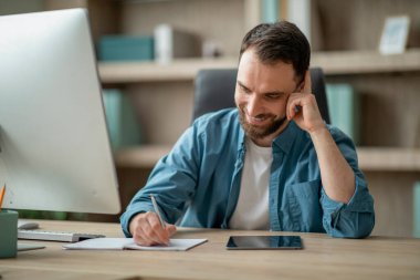 Yakışıklı genç iş adamı cep telefonuyla konuşuyor ve ofiste çalışırken notlar alıyor, kalemini tutuyor ve not alıyor, erkek girişimci masasında oturuyor ve telefon görüşmesinin keyfini çıkarıyor, boş zaman aralığının tadını çıkarıyor.