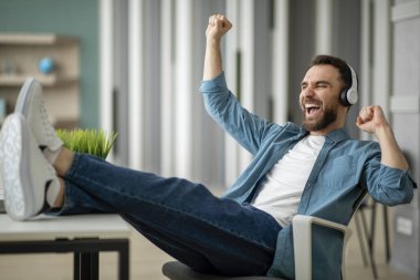 Break In Work. Ofiste müzik dinleyen mutlu genç adam, kablosuz kulaklıklı neşeli erkek çalışan favori şarkıların ve dansların tadını çıkaran, bacaklarını masaya koyan,