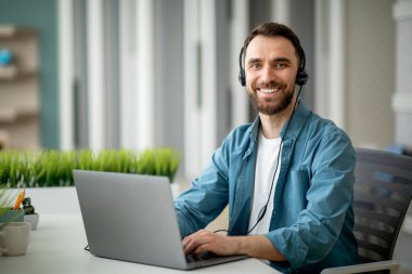Acil yardım hattı operatörü. Ofiste çalışan yakışıklı müşteri destek müdürü portresi, masadaki dizüstü bilgisayarla çalışan, kameraya bakıp gülümseyen kulaklıklı erkek.