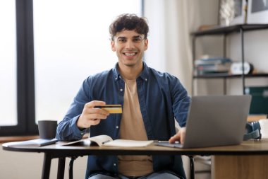 E-Ticaret Teklifi. Orta Doğulu neşeli iş adamı, kredi kartı ve laptop alışverişi kullanıyor. Modern ofiste işyerinde oturuyor. Neşeli Girişimci Ödemeleri Çevrimiçi Bankacılık yoluyla Yapıyor