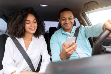 Genç çift birlikte araba yolculuğuna çıkıyor, telefonla mobil uygulama kullanıyor, ön otomobil koltuklarında oturuyor, araç panelinden manzara seyrediyor. Sürücü cep telefonunu otomatik olarak karısına gösteriyor