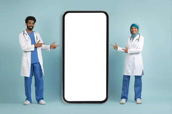 穿着制服的穆斯林医生站在蓝色工作室的背景下在大空白智能手机屏幕上微笑 阿拉伯医生展示医疗广告的复制空间 — 图库照片