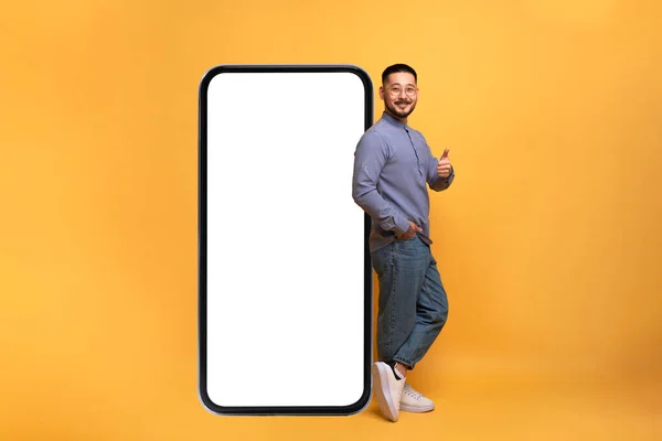 モバイルオファー 興奮したアジアの男は 空白の画面で大きなスマートフォンにもたれ ジェスチャーを表示します 100万人の男は素晴らしい新しいアプリやウェブサイトをお勧めします 黄色の背景 モックアップ — ストック写真