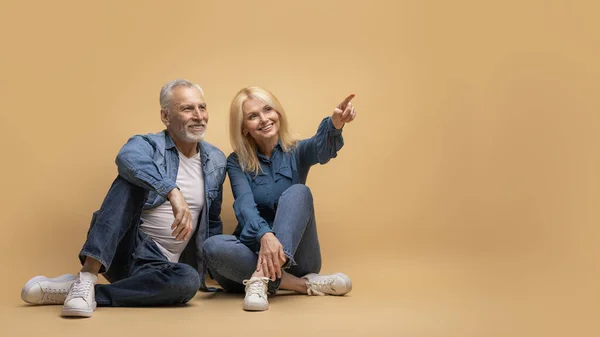 Happy positive elderly couple grey-haired man and blonde woman wearing casual outfit sitting together on floor over beige background, pointing at copy space, showing nice deal, panorama