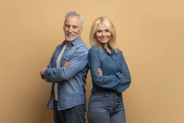 stock image Partnership, teamwork concept. Cheerful attractive confident senior man and woman wearing casual denim outfit standing back to back with arms crossed over beige studio background, smiling at camera