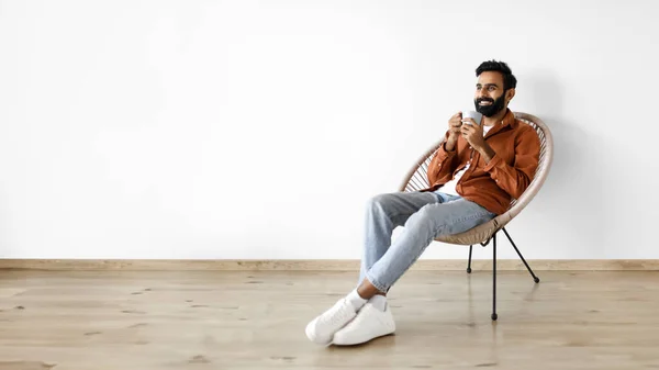 Cheerful indian man drinking aromatic coffee while chilling in comfy armchair on white wall background, at modern home interior. Bearded guy having hot beverage, relaxing during his break. Panorama