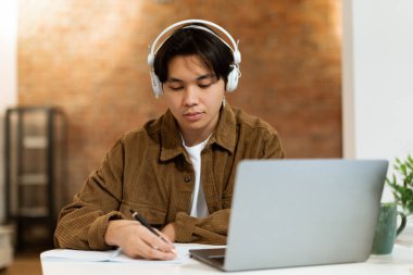 Ciddi Asyalı öğrenci, kulaklıklı, dizüstü bilgisayarının yanında notlarını yazan, web sörfü yapan ve ödev yapan ev içi çalışma odasındaki çalışma masasında oturan. Eğitim ve Evde Eğitim Konsepti