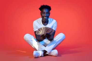 Hediye, nakit para, piyango. Mutlu, yakışıklı, yakışıklı, sıradan giyinmiş beyaz adam kırmızı arka planda oturuyor, elinde nakit para banknotları, fotokopi alanı var.