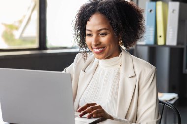 Kadınlar iş başında. Resmi kıyafet yöneticisi olan neşeli genç siyahi kadın, CEO bilgisayar dizüstü bilgisayarının önünde oturuyor, klavyeye oturuyor ve gülümsüyor, iyi haberler var, modern ofis içi