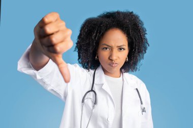 Üzgün, kızgın, kıvırcık saçlı, beyaz önlüklü Amerikalı kadın doktor terapist mavi arka planda, stüdyoda izole edilmiş bir şekilde işaret veriyor. Sorunlar, sağlık hizmetleri ve hastalıklar hakkında kötü eleştiriler