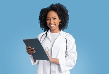 Pozitif genç Afro-Amerikan kıvırcık kadın terapist beyaz önlüklü, tablet üzerinde sohbet eden, mavi arka plan, stüdyo. Uzaktan sağlık hizmeti, uygulama tıp hizmeti, sınav