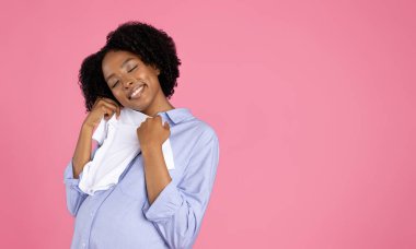 Milenyum Afro-Amerikan kıvırcık saçlı hamile kadın günlük bebek kıyafetleri içinde, pembe stüdyo arka planında izole edilmiş gelecekteki anneliğin tadını çıkarıyor. Alışveriş, çocuk almak, çocuk sahibi olmak.