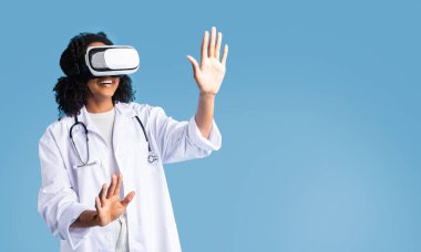 Beyaz önlüklü ve VR gözlüklü mutlu Afro-Amerikan kıvırcık kadın mavi arka planda, stüdyoda izole edilmiş ellerle boşluğa dokunuyor. Teknoloji, bilim, tıp ve sağlık hizmetleriyle tedavi