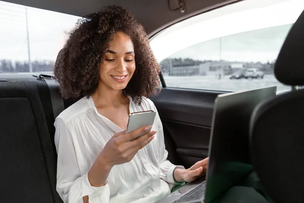 いつも連絡を取り合ってる 車の後部座席に座ってノートパソコンを使用して 携帯電話でテキストメッセージを送信し 自動車旅行中にオンラインで作業し 通信している陽気なアラビア語のビジネス女性 事業譲渡 — ストック写真